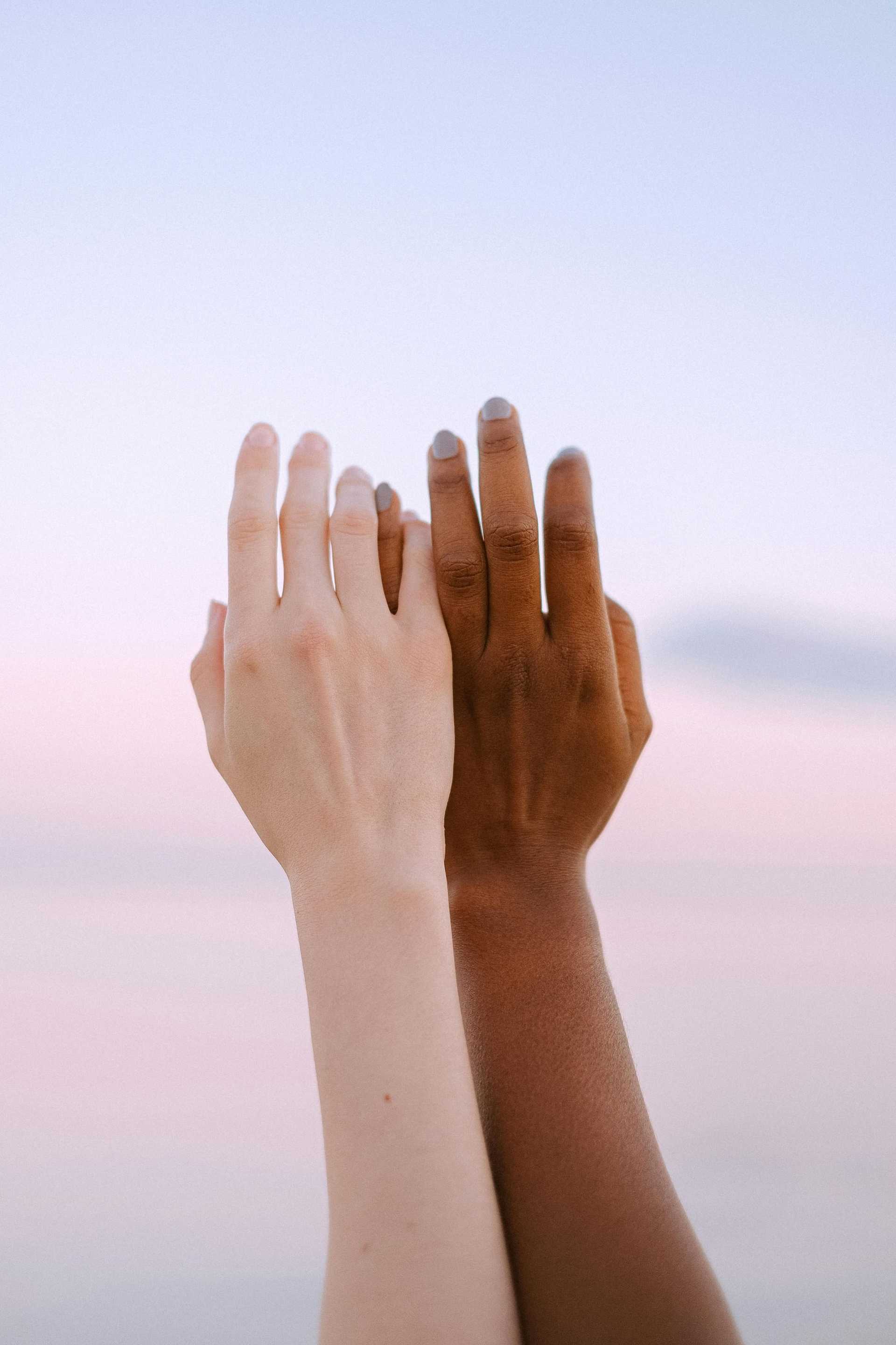 Two hands of different skin tones reaching upwards against a serene sky.